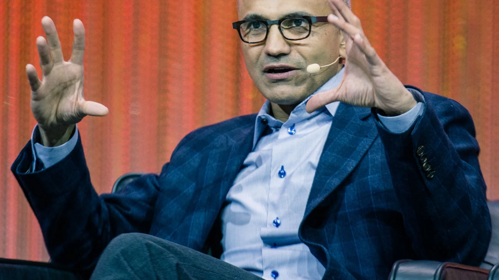 A bald man wearing glasses, a blue suit jacket, and a light-blue shirt sits in a chair with his legs crossed.