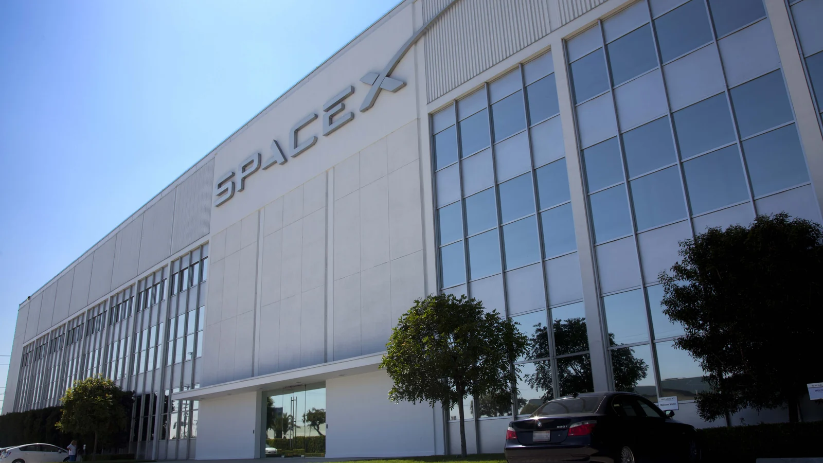 A large modern building with a white metal-and-panel facade, extensive grid-patterned glass windows, and the raised word “SPACEX” on its upper wall.