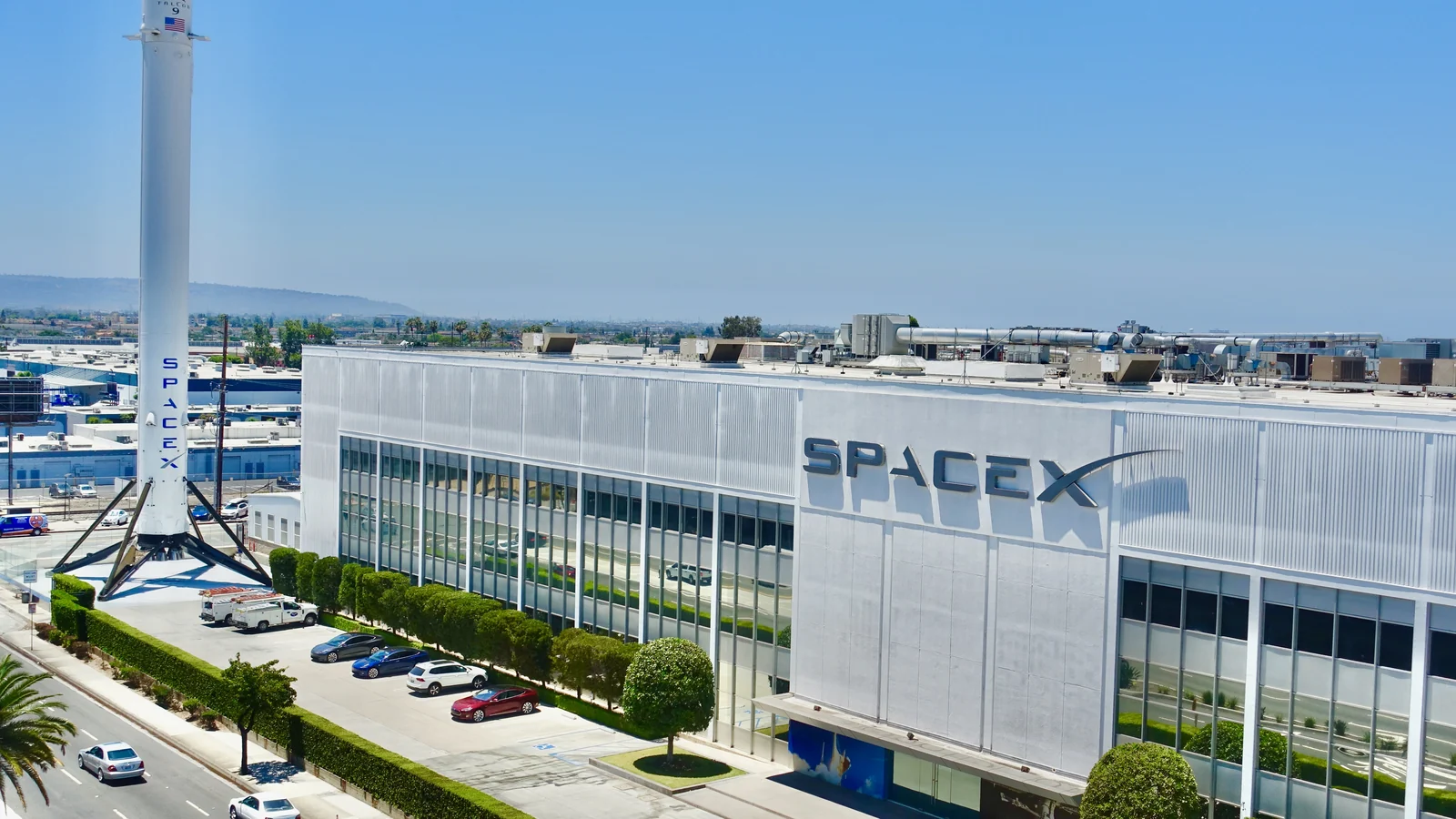 A large white industrial building with extensive glass windows and the word “SPACEX” displayed on its facade.