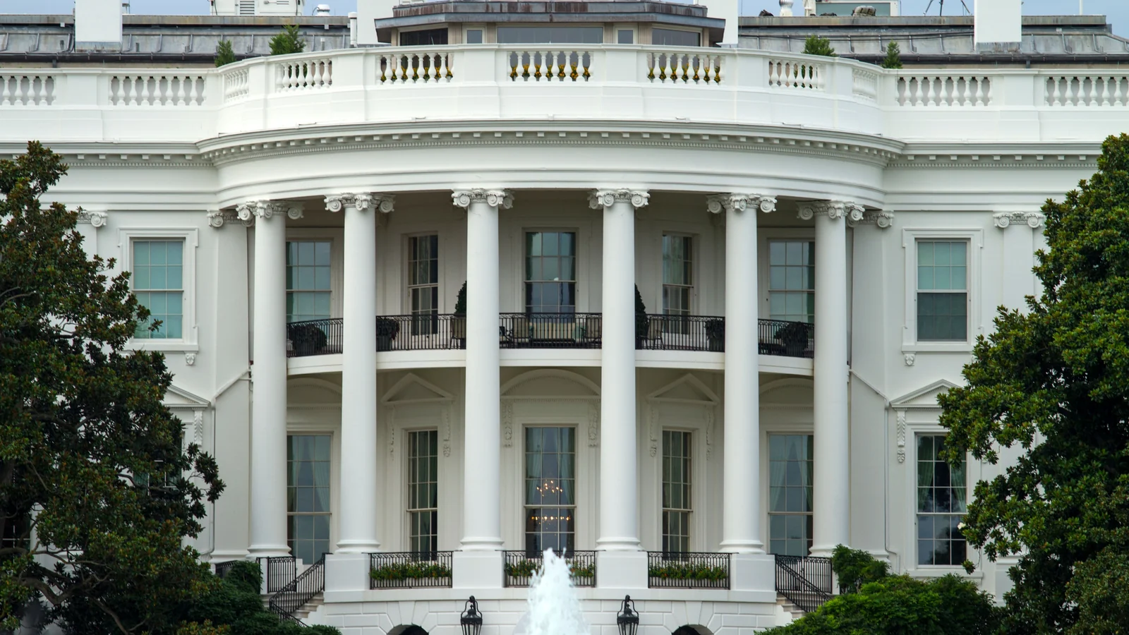 A large white neoclassical building is visible, featuring a curved portico with tall columns, ornate capitals, balustrades, tall multi-pane windows, rooftop chimneys and…