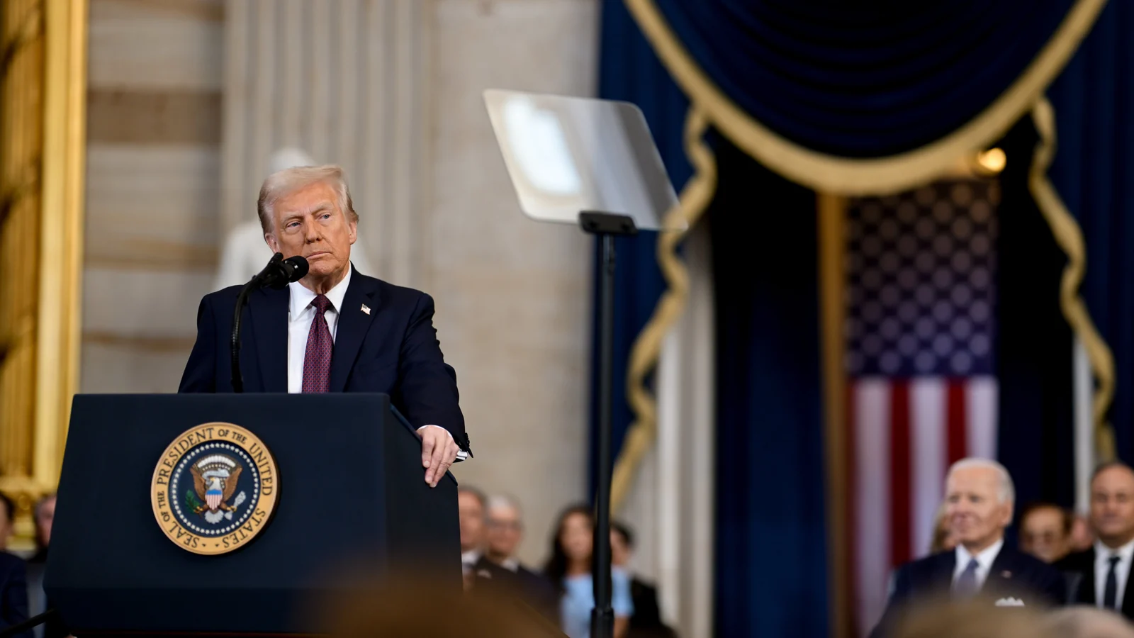 An older man in a dark suit and red tie stands behind a lectern bearing a circular presidential seal, speaking into a microphone.