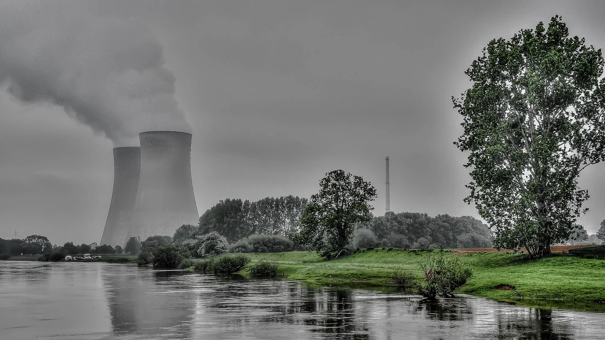 A riverside landscape with two large hyperboloid concrete cooling towers in the background, one partly behind the other, and a tall narrow industrial chimney nearby.